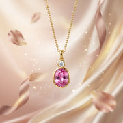 Gold pendant necklace featuring an oval pink gemstone and a small round clear accent stone, displayed against a soft pink satin background with floating petals and sparkles.
